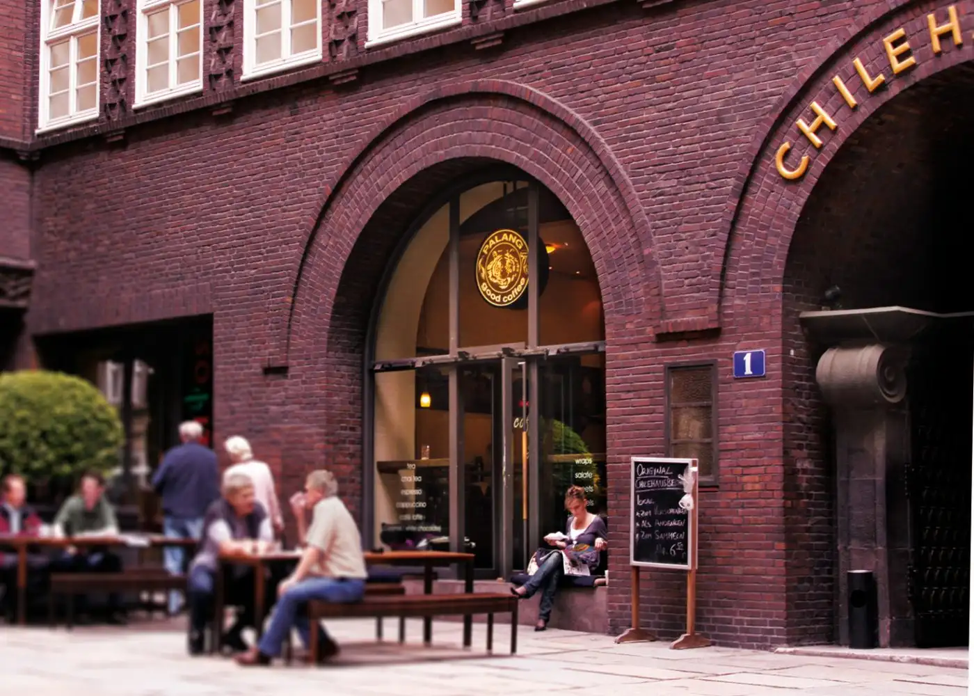 Eingang von PALANG good coffee im Chilehaus: Gäste an Tischen vor dem Backstein-Bogen, das Logo im Fenster